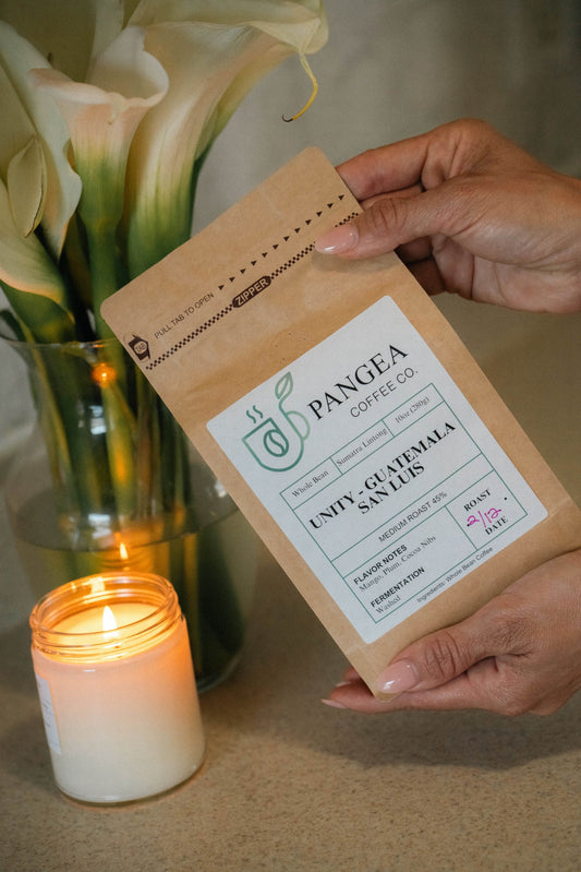 Pangea Coffee’s Unity Medium Roast bag placed beside a softly lit candle – warm, calming ambiance highlighting the comforting nature of this Roaster’s Choice blend.